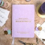 Mein kleines Hochzeitsbuch Hochzeitsplaner Wedding Planner Lila - Flatlay-Ansicht des Hochzeitsplaners auf einem braunen, dekorierten Tisch mit Accessoires, Wachssiegel, Hochzeitseinladung, Kerze