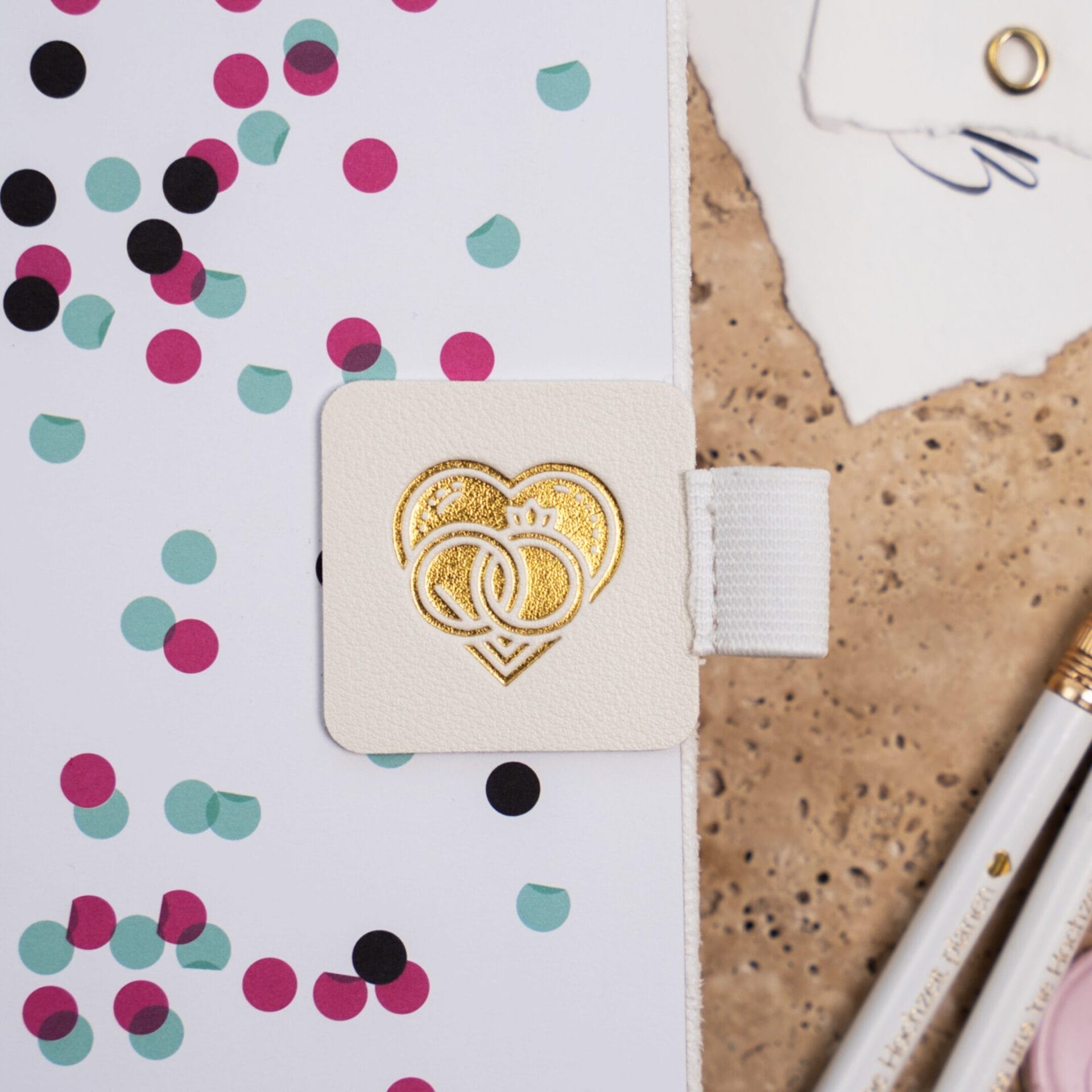 Stifthalter Stiftehalter Stiftschlaufe Pen Loop Hochzeitsplaner Weiß - Befestigt auf einem Hochzeitsbuch. Brauner Hintergrund mit Hochzeitsplanung zu sehen. Zwei weiße Bleistifte mit goldener Gravur
